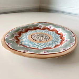 Pu&ograve; includere: Un piatto decorativo in ceramica con un design dipinto a mano. Il piatto presenta un bordo marrone chiaro, un'area centrale con motivi blu e bianchi e un bordo con accenti verdi, marroni e viola. Il piatto &egrave; rotondo e ha una forma leggermente irregolare.