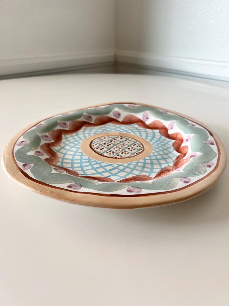 Pu&ograve; includere: Un piatto decorativo in ceramica con un design dipinto a mano. Il piatto presenta un bordo marrone chiaro, un'area centrale con motivi blu e bianchi e un bordo con accenti verdi, marroni e viola. Il piatto &egrave; rotondo e ha una forma leggermente irregolare.