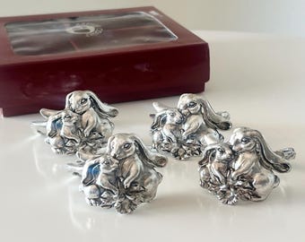 May include: Set of five silver-toned bunny napkin rings. Each ring features a mother rabbit and a baby rabbit. A burgundy box with a clear lid is in the background.