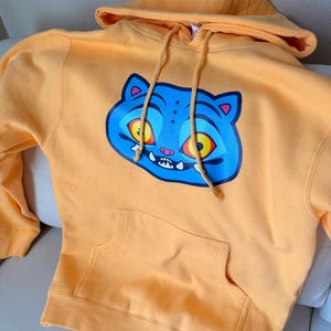 May include: A light orange hoodie with a large blue cat face graphic on the front. The cat has yellow eyes, a pink mouth, and white teeth. The hoodie has a drawstring hood and a front pocket.