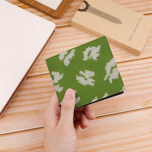 May include: A green wallet with a white camouflage pattern is held in a hand. The wallet is rectangular and appears to be made of a textured material. The background includes a wooden surface and a box.