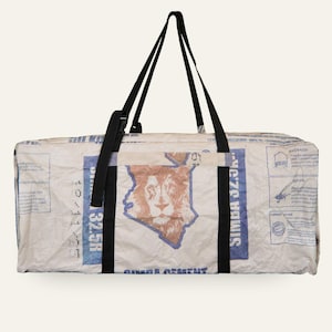 Könnte beinhalten: Eine rechteckige Reisetasche aus wiederverwertetem, cremefarbenem Webmaterial mit blauem Text und einer braunen Löwengrafik innerhalb eines Kartenumrisses. Die Tasche verfügt über schwarze Nylongurte und einen schwarzen Kunststoffverschluss.