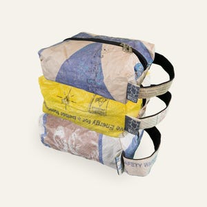 Könnte beinhalten: Ein Stapel aus drei rechteckigen Taschen aus wiederverwertetem Webmaterial. Die oberste Tasche ist blau und beige, die mittlere ist gelb mit dem Text "Energy for a better tomorrow" und die untere ist braun und weiß. Jede hat einen schwarzen Reißverschluss.