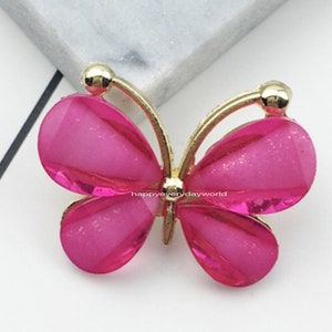 Puede incluir: Un adorno decorativo con forma de mariposa con un marco dorado y alas rosas brillantes facetadas. El tamaño es de 2,2 x 2,8 mm. La mariposa está sobre un fondo blanco y gris.