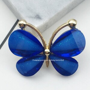 Puede incluir: Un adorno de resina azul en forma de mariposa con detalles dorados. El adorno mide 2,2 x 2,8 mm.