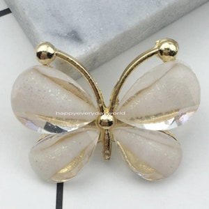 Puede incluir: Un adorno de resina en forma de mariposa blanca con detalles dorados. La mariposa mide 2,2 x 2,8 mm.