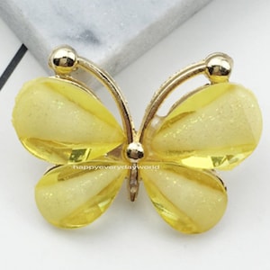 Puede incluir: Un adorno de resina de color amarillo en forma de mariposa con detalles en dorado. El adorno mide 2,2 x 2,8 mm.