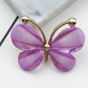 Puede incluir: Adorno de resina en forma de mariposa púrpura con detalles dorados. La mariposa mide 2,2 x 2,8 mm.