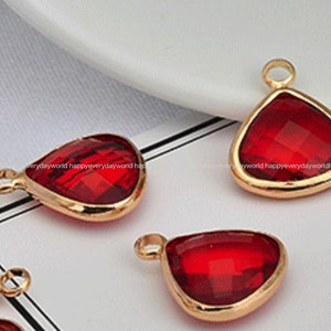 May include: Close-up of several red teardrop-shaped pendants with gold-colored metal frames. Each pendant has a faceted, jewel-like appearance and a small loop at the top for attaching to a chain or other jewelry component. The pendants are displayed on a white surface.