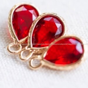 May include: Three teardrop-shaped red glass charms with gold-toned metal settings. The charms are arranged in a row, with the largest charm at the back and the smallest charm at the front.