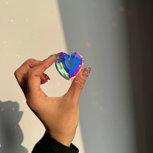 Op de afbeelding: Een hartvormige kristallen hanger met een iriserende afwerking die tinten blauw, paars en groen reflecteert. Het gefacetteerde oppervlak vangt het licht en creëert een prismatisch effect met een klein gaatje aan de bovenkant voor bevestiging.