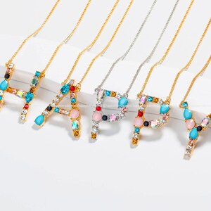 May include: Gold-toned necklaces spelling "HAPPY" with colorful gemstone letter pendants. Each letter is adorned with a variety of gems in shades of blue, pink, and clear, creating a sparkling effect. The necklaces are displayed on a white surface.
