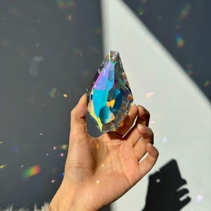 Könnte beinhalten: Ein facettiertes, tropfenförmiges Kristallprisma, das ein lebendiges, irisierendes Regenbogenspektrum zeigt, wenn Licht hindurchfällt. Das klare Glasobjekt weist mehrere geometrische Winkel und scharfe Kanten auf, die das Licht brechen.