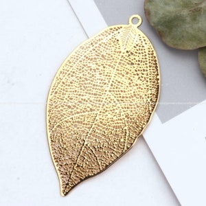 Puede incluir: Un colgante de hoja de metal de color dorado con un diseño texturizado detallado. La hoja tiene forma ovalada y tiene un agujero en la parte superior para colgar.