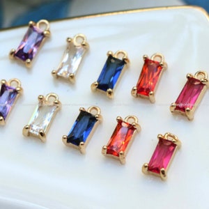 Pode incluir: Uma coleção de pingentes retangulares de pedras preciosas em várias cores, incluindo roxo, transparente, azul, vermelho e rosa. Cada pingente tem uma moldura de metal dourado e um pequeno laço na parte superior.