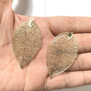 Puede incluir: Dos abalorios de metal en forma de hoja de color dorado con un diseño delicado e intrincado. Los abalorios tienen un agujero en la parte superior para sujetarlos a joyas u otras manualidades.