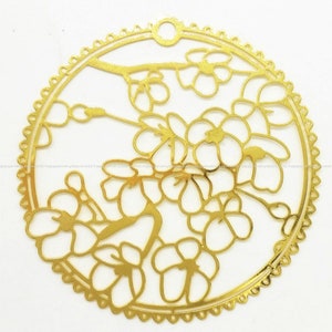 May include: A gold-colored, circular metal cutout featuring a floral design. The cutout showcases delicate flowers and branches within a scalloped border. A small hole is at the top for hanging or attachment.