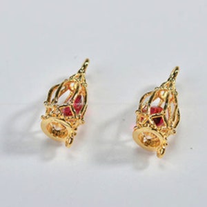 Puede incluir: Dos colgantes de jaula de color dorado con detalles de cristal rojo.