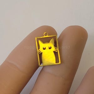 Könnte beinhalten: Goldfarbener Katzenanhänger mit rechteckigem Rahmen. Die Katze hat ein einfaches, stilisiertes Design mit zwei kleinen Augen und einer kleinen Nase. Der Anhänger hat oben eine Schlaufe zum Befestigen an einer Kette oder anderem Schmuck.