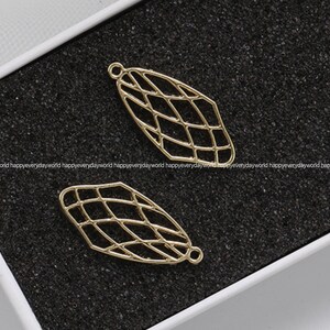 May include: Two gold-colored, openwork leaf-shaped charms with a geometric grid pattern. Each charm has a small loop at the top for attaching to jewelry. The charms are displayed on a dark, textured surface.