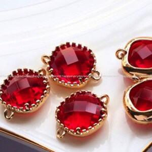 May include: Close-up of several red gemstone charms with gold-coloured settings. The gems have a faceted cut, and the settings have small loops for attaching to jewellery. The charms are arranged on a white surface.
