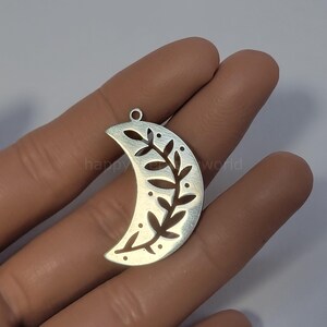 Op de afbeelding: Zilverkleurige maanvormige hanger met een uitgesneden bladontwerp en kleine stipaccenten. De hanger heeft een kleine lus aan de bovenkant om op te hangen. De tekst "happy world" is gedeeltelijk zichtbaar.