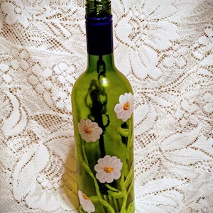 May include: A green glass wine bottle decorated with white daisy flowers and green stems. The bottle has a blue neck and a metal ring attached to the top. The flowers have yellow centers.