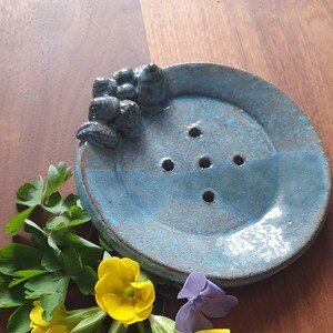 Könnte beinhalten: Eine blaue Keramik-Seifenschale mit einem dekorativen Design aus drei kleinen, dunkelblauen, abgerundeten Formen. Die Schale hat fünf Abflusslöcher und steht auf einer Holzoberfläche mit gelben und blauen Blumen.