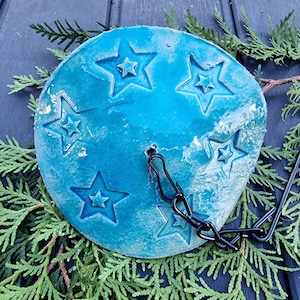 Könnte beinhalten: Ein rundes, türkisfarbenes Keramikstück mit geprägten Sternmustern. Eine schwarze Metallkette und ein Haken sind angebracht. Das Stück ist von grünem Laub umgeben und bildet einen dekorativen Akzent.