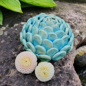 Könnte beinhalten: Ein blaues dekoratives Keramikobjekt mit strukturierter Oberfläche, das einem Steinhaufen ähnelt, liegt auf einem Felsen. Zwei weiße und rosa Blumen befinden sich neben dem Keramikobjekt. Der Hintergrund umfasst grüne Blätter und eine natürliche Steinoberfläche.