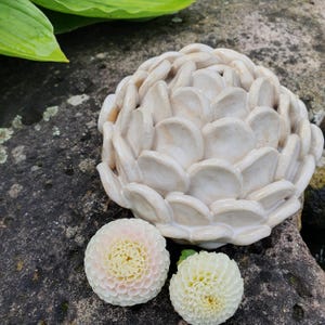 Könnte beinhalten: Eine weiße Keramikskulptur einer Blume mit einem geschichteten Design. Die Skulptur steht auf einem Stein mit zwei weißen Blumen.
