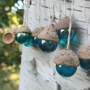Könnte beinhalten: Dekorative, hängende Ornamente in Form von Eicheln. Jedes Ornament hat eine braune Kappe und einen teal-farbenen, irisierenden Glaskörper. Die Ornamente sind an dünnen Schnüren aufgehängt und erzeugen eine verspielte, von der Natur inspirierte Ästhetik.