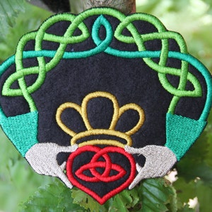 Könnte beinhalten: Gestickter Aufnäher mit einem grünen keltischen Knotendesign, einer goldenen Krone und einem roten Herzen mit einem keltischen Knoten darin. Der Aufnäher ist auf schwarzem Hintergrund.