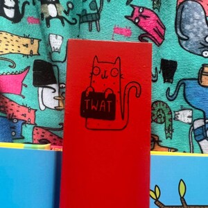May include: A red wooden bookmark with a black and white illustration of a cat holding a sign that says "TWAT".