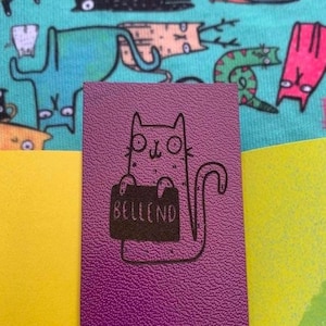 May include: A purple leather notebook with a black and white illustration of a cat holding a sign that says "Belleend".