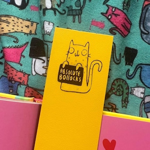 May include: A yellow bookmark featuring a cartoon cat holding a sign that reads "ABSOLUTE BOLLOCKS".
