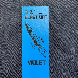 May include: A blue bookmark with a black rocket ship illustration and the text "3...2...1... BLAST OFF" and "VIOLET" printed on it.