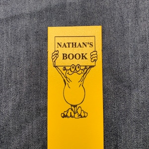 May include: A yellow bookmark featuring a cartoon character holding a sign that reads "NATHAN'S BOOK".