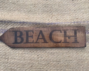 May include: A wooden sign with a pointed end, painted brown, with the word "BEACH" in black lettering.