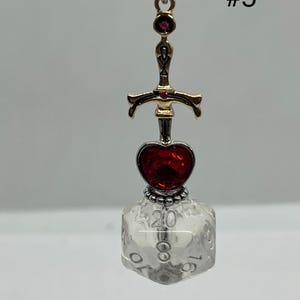 Peut inclure: Un porte-cl&eacute;s compos&eacute; d'une breloque &eacute;p&eacute;e dor&eacute;e avec de petits accents rouges, une gemme rouge en forme de c&oelig;ur et un d&eacute; &agrave; vingt faces transparent avec des chiffres noirs. Le chiffre 20 est visible en haut du d&eacute;. Le texte #5 est visible en haut &agrave; droite.
