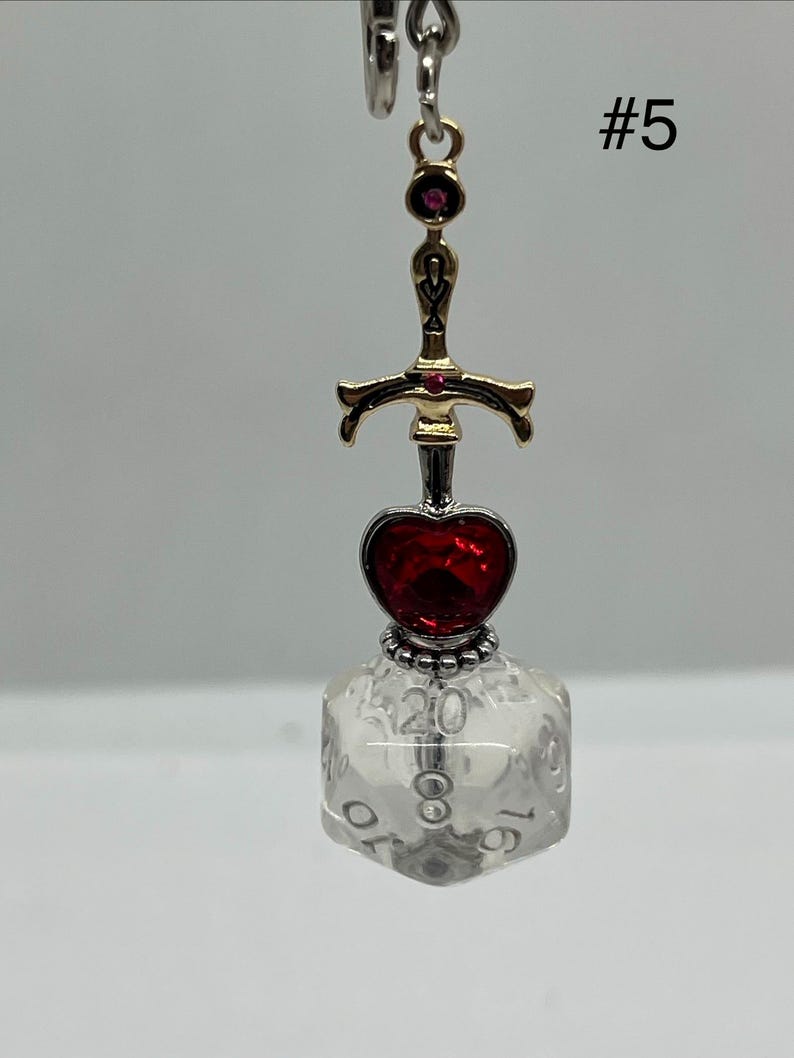 Peut inclure: Un porte-cl&eacute;s compos&eacute; d'une breloque &eacute;p&eacute;e dor&eacute;e avec de petits accents rouges, une gemme rouge en forme de c&oelig;ur et un d&eacute; &agrave; vingt faces transparent avec des chiffres noirs. Le chiffre 20 est visible en haut du d&eacute;. Le texte #5 est visible en haut &agrave; droite.