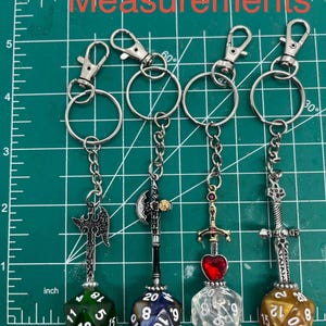 Peut inclure: Quatre porte-cl&eacute;s en m&eacute;tal avec des armes fantastiques miniatures et des d&eacute;s &agrave; vingt faces. Les armes incluent deux haches et deux &eacute;p&eacute;es, dont une avec un c&oelig;ur rouge. Les d&eacute;s sont verts, bleus, transparents et dor&eacute;s. Une r&egrave;gle indique moins de 13 cm.