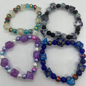 May include: Four beaded bracelets with dice-shaped beads and round beads in various colors. The bracelets feature clear, purple, blue, and black dice with gold numbering. The round beads include pearls and colorful glass beads.
