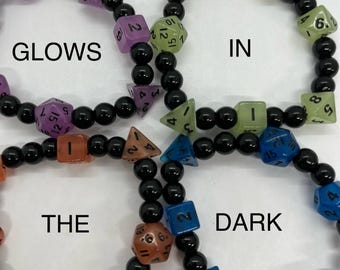 Peut inclure: Quatre bracelets circulaires composés de perles noires alternées avec des dés polyédriques violets, vert clair, orange et bleus. Les dés portent des chiffres noirs. Le texte GLOWS IN THE DARK est superposé sur l'image des quatre bracelets.