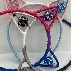 May include: A collection of cat ear headbands wrapped in satin ribbon in shades of pink, white, blue, purple, and black. Each headband features integrated 20-sided dice in matching or contrasting colors, accented with small decorative beads.