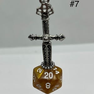 Peut inclure: Un pendentif &eacute;p&eacute;e en m&eacute;tal argent&eacute; avec une garde textur&eacute;e, attach&eacute; &agrave; un d&eacute; &agrave; vingt faces de couleur ambre avec des chiffres grav&eacute;s en blanc. Le chiffre 20 est clairement visible sur la face avant. Le texte #7 appara&icirc;t dans le coin sup&eacute;rieur droit.