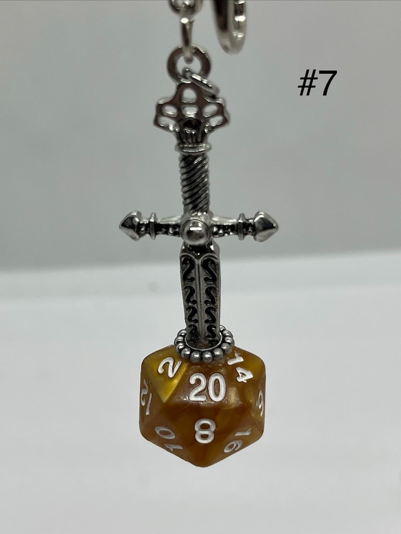 Peut inclure: Un pendentif &eacute;p&eacute;e en m&eacute;tal argent&eacute; avec une garde textur&eacute;e, attach&eacute; &agrave; un d&eacute; &agrave; vingt faces de couleur ambre avec des chiffres grav&eacute;s en blanc. Le chiffre 20 est clairement visible sur la face avant. Le texte #7 appara&icirc;t dans le coin sup&eacute;rieur droit.