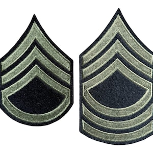 May include: Two green and black military rank insignia patches. The patches are shaped like a chevron with three stripes. The stripes are outlined in a darker green colour.