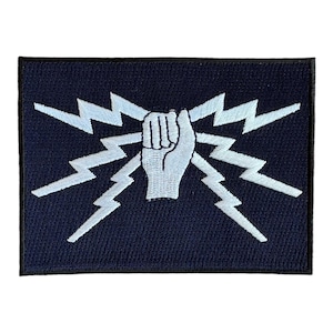 May include: A white embroidered patch on a dark blue background. The patch features a white fist with lightning bolts radiating from it.