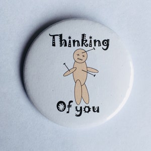 May include: A white button with a black and brown illustration of a voodoo doll with the text "Thinking of you" on it.
